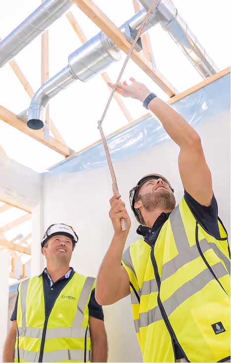 Two construction workers wearing yellow reflective vests and white helmets inspecting ductwork in a building under construction.
