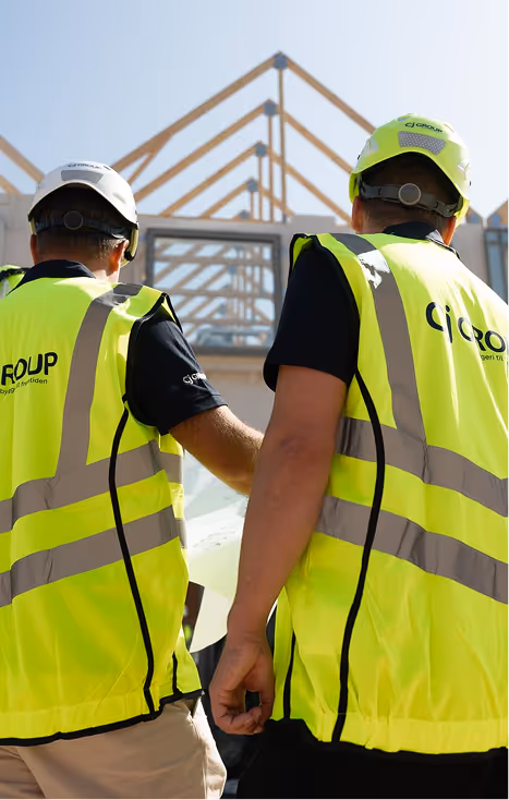 Two construction workers wearing high-visibility vests and helmets reviewing building plans at a construction site with wooden roof frames in the background.