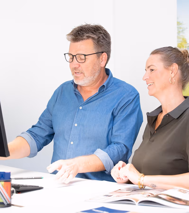 Man in a blue shirt and glasses discussing something on a computer with a smiling woman in a dark polo shirt at a white desk.