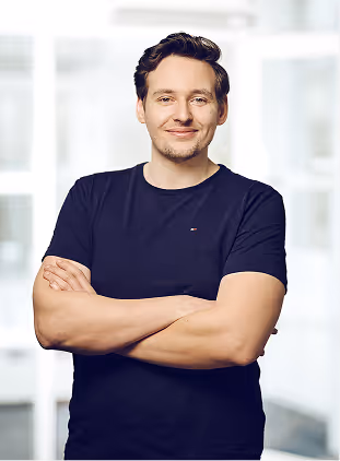Smiling man with short brown hair wearing a black t-shirt, standing with arms crossed in a bright indoor setting.