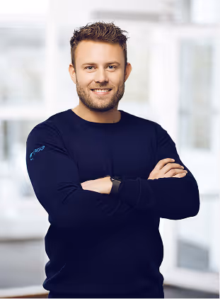 Smiling man with short curly hair wearing a dark sweater and smartwatch, standing with arms crossed in a bright modern office.