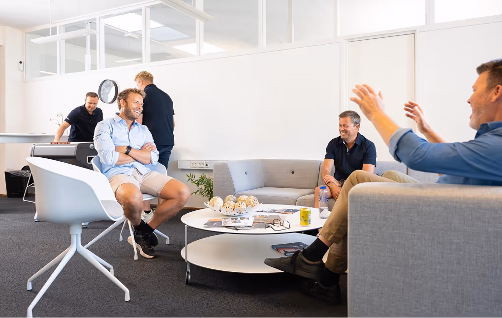 Five men casually interacting in a modern office lounge with gray sofas, a white round table, and a foosball table in the background.