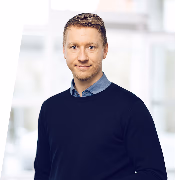 Smiling man with short light brown hair wearing a dark sweater over a blue collared shirt standing indoors.