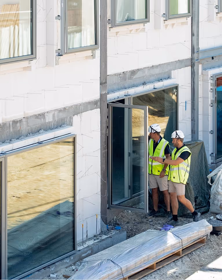 Two construction workers in yellow safety vests and white helmets inspecting a glass door on a building under construction.