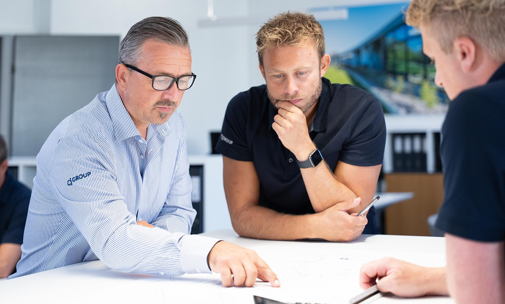 Three men in a meeting room leaning over a table reviewing documents, one pointing to the papers and another holding a pen.