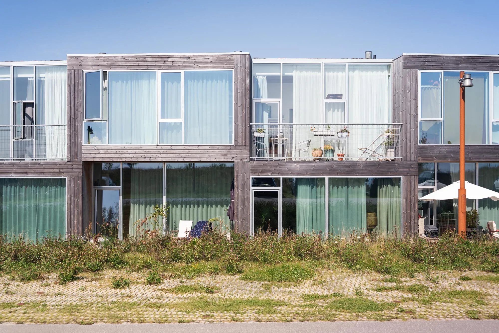Moderne rækkehus med træfacade, store vinduer, altan med havemøbler og planter under klar blå himmel.