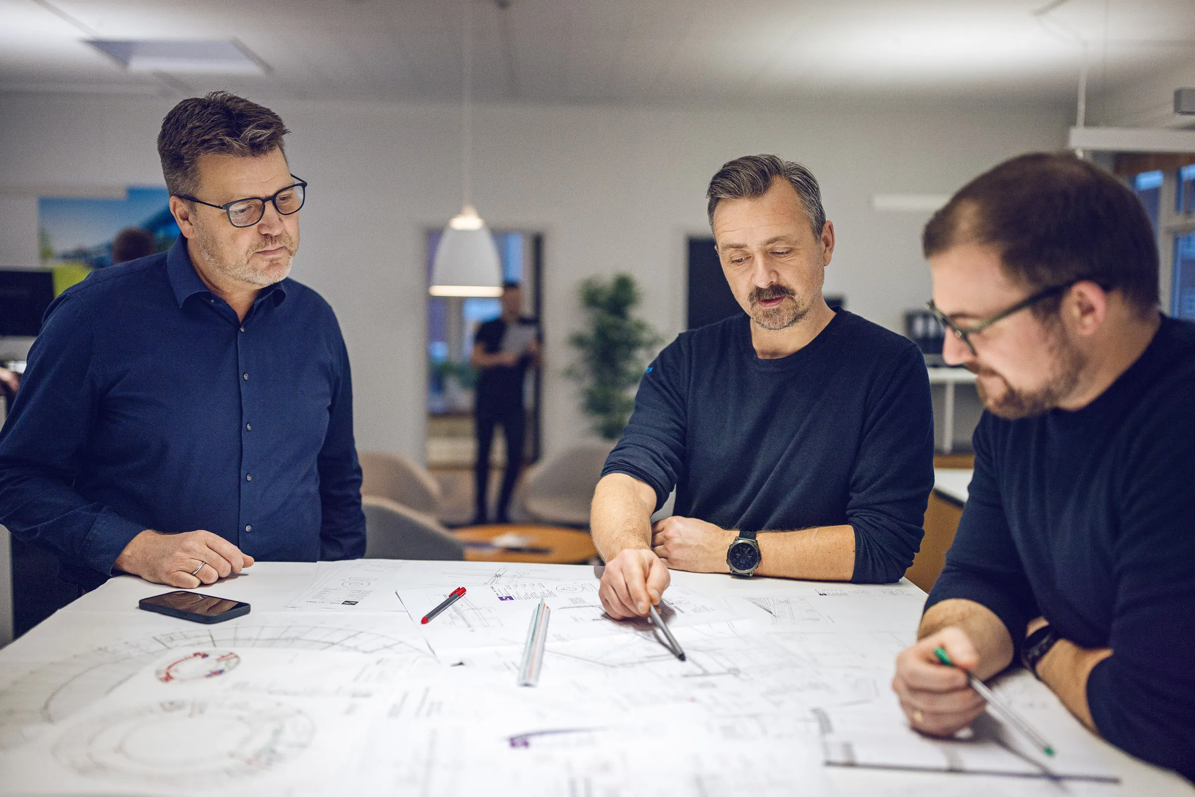 Tre mænd diskuterer og gennemgår tekniske tegninger på et bord i et kontormiljø.