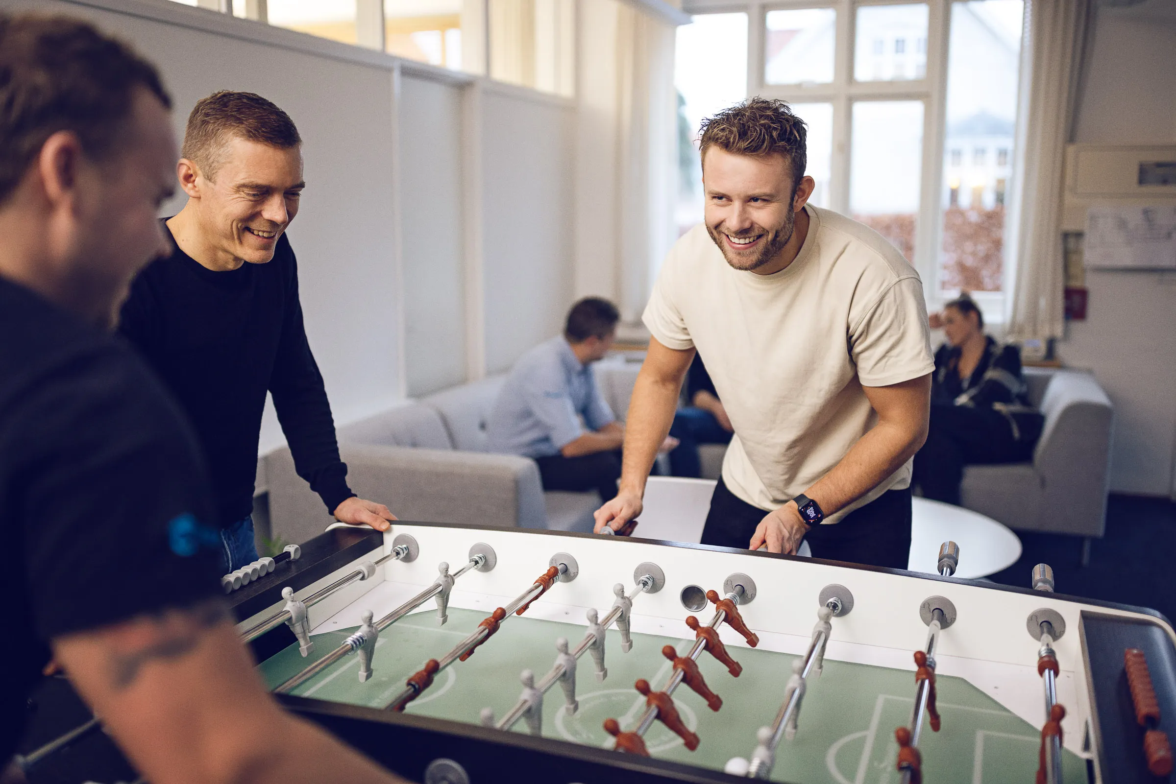 Tre mænd smilende spiller bordfodbold i et lyst opholdsområde, mens to personer sidder i baggrunden.