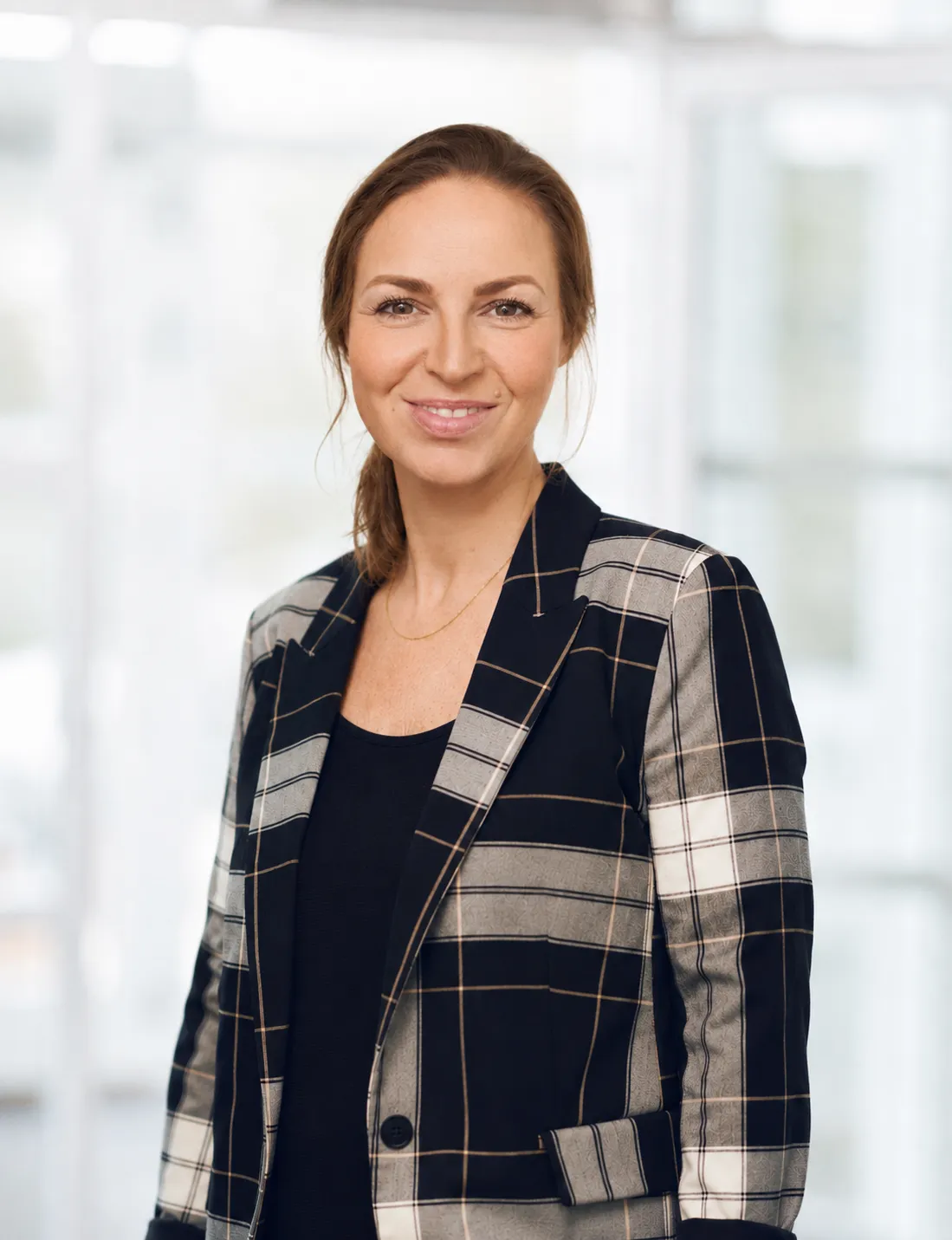 Smilende kvinde iført sort top og ternet blazer foran lys og udvisket baggrund.