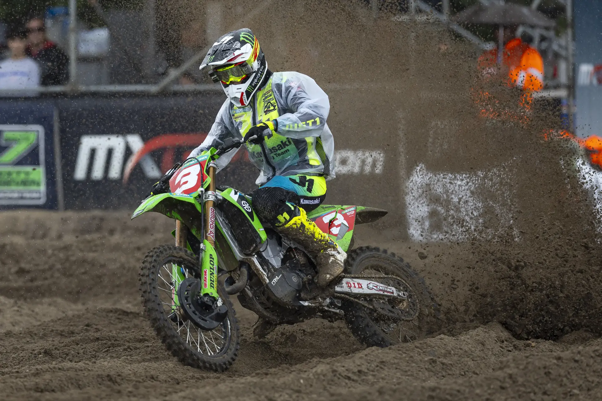 Motocross rider in yellow and black gear on a green Kawasaki bike kicking up dirt on a muddy track.