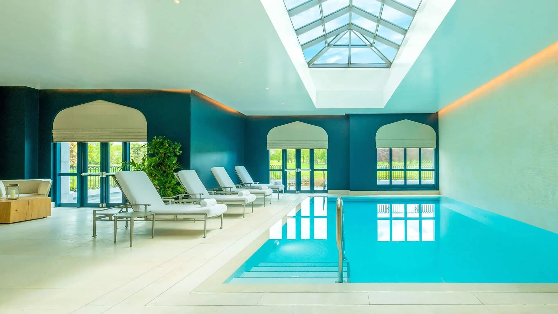 Adare Manor Resort. Indoor swimming pool with blue water, white lounge chairs, large windows, and a skylight ceiling.