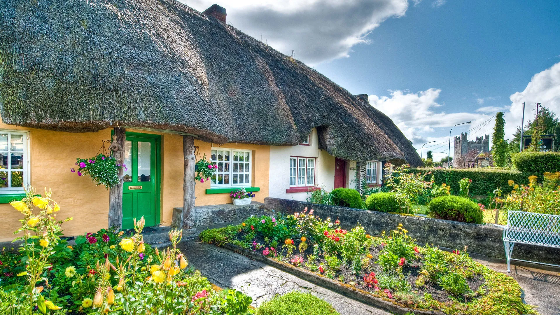 Adare Village, County Limerick. Thatched cottages with colorful flower gardens and green doors under a blue sky.