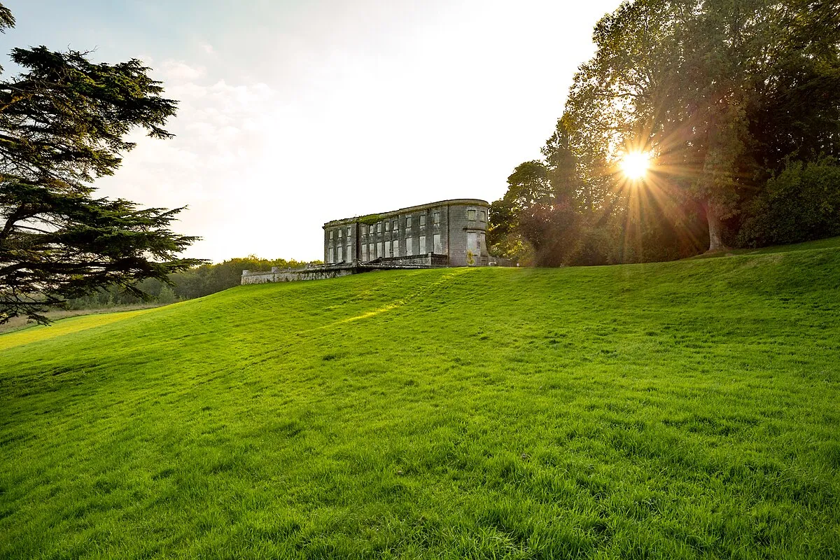 Curraghchase. Sunlight shining through trees beside a historic stone building on a green hill.