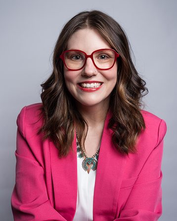 Smiling woman with wavy brown hair wearing red glasses, a pink blazer, and a turquoise necklace on a light gray background.
