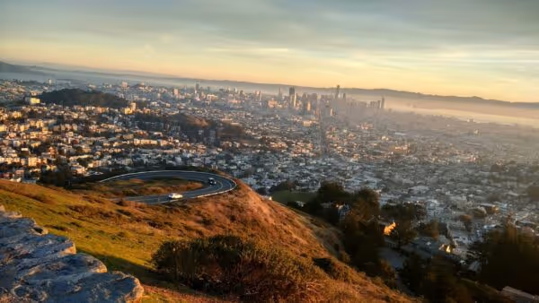 Sunset view of a city skyline with a winding road and grassy hill in the foreground.