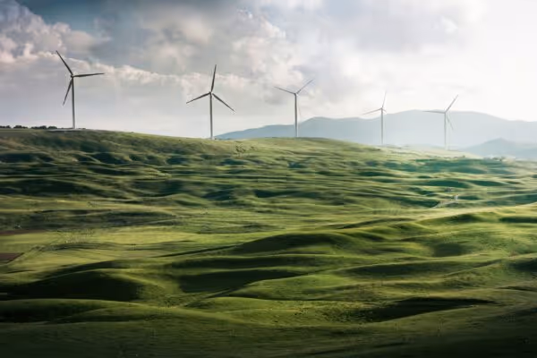 Wind turbines standing on rolling green hills under a cloudy sky with mountains in the distance.