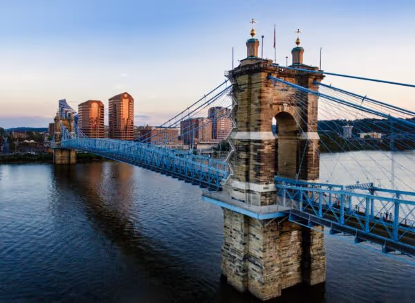 Suspension bridge spanning a calm river with city buildings in the background at sunset.