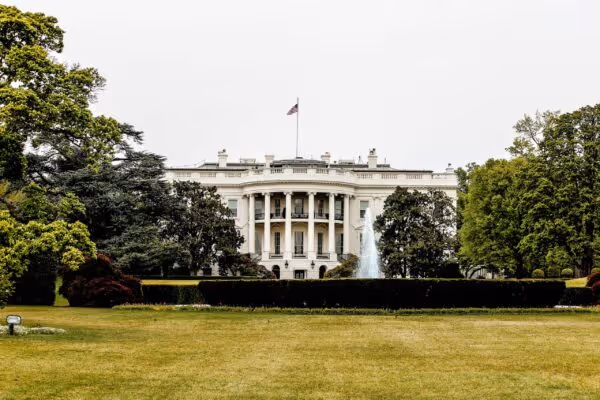 Front view of the White House with a lawn, bushes, and trees surrounding it under a gray sky.