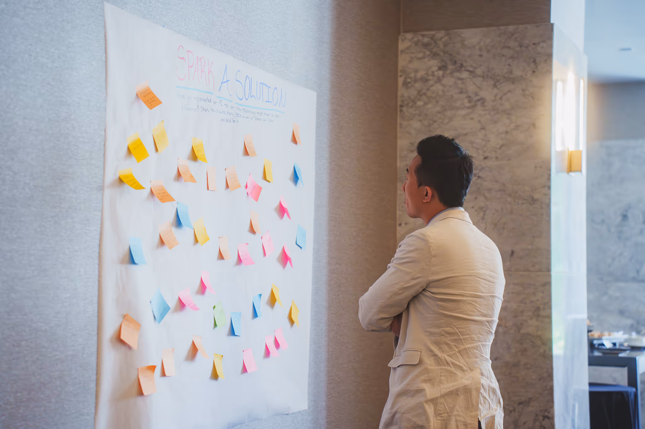 Man with arms crossed reviewing colorful sticky notes on a "Spark a Solution" brainstorming board at a workshop
