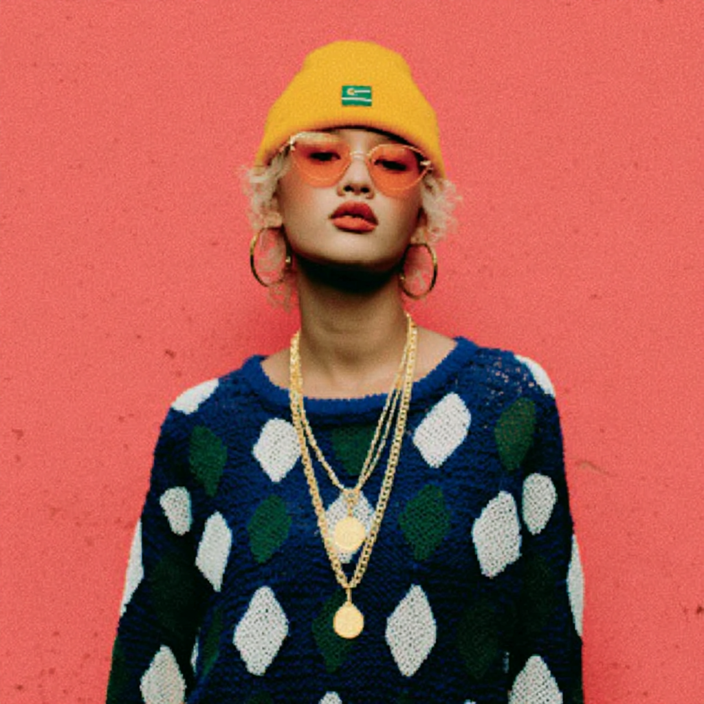 Person with curly blonde hair wearing a yellow beanie, orange sunglasses, large hoop earrings, layered gold necklaces, and a blue sweater with green and white diamond patterns, standing against a red background.