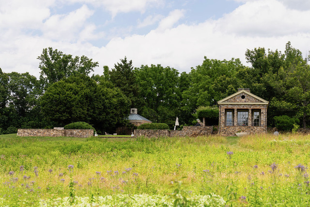 Stone building and surrounding stone walls set behind a field of wildflowers with dense green trees in the background under a partly cloudy sky.