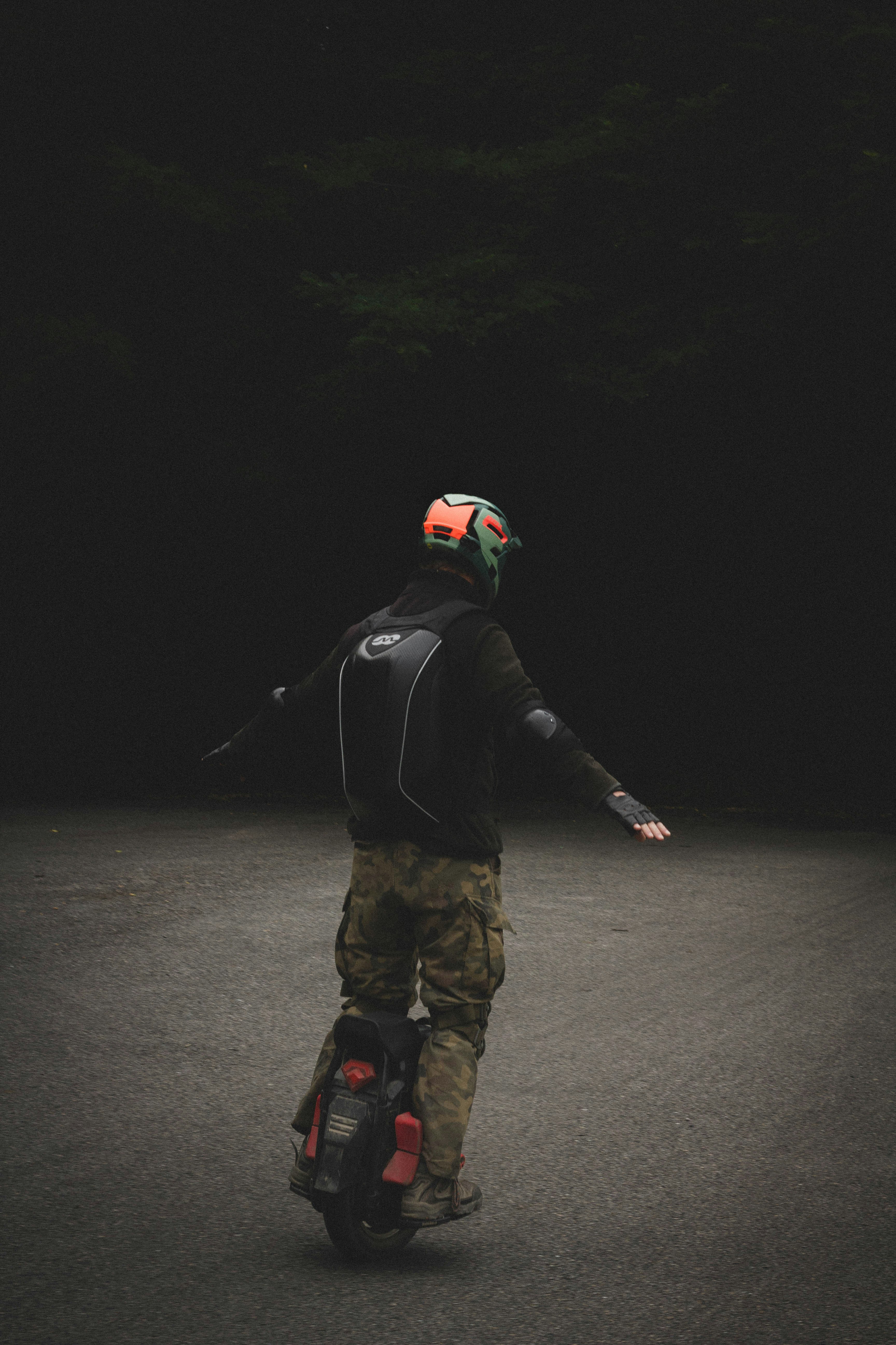 Person in camo pants and helmet riding a single-wheeled electric unicycle on a dark road.