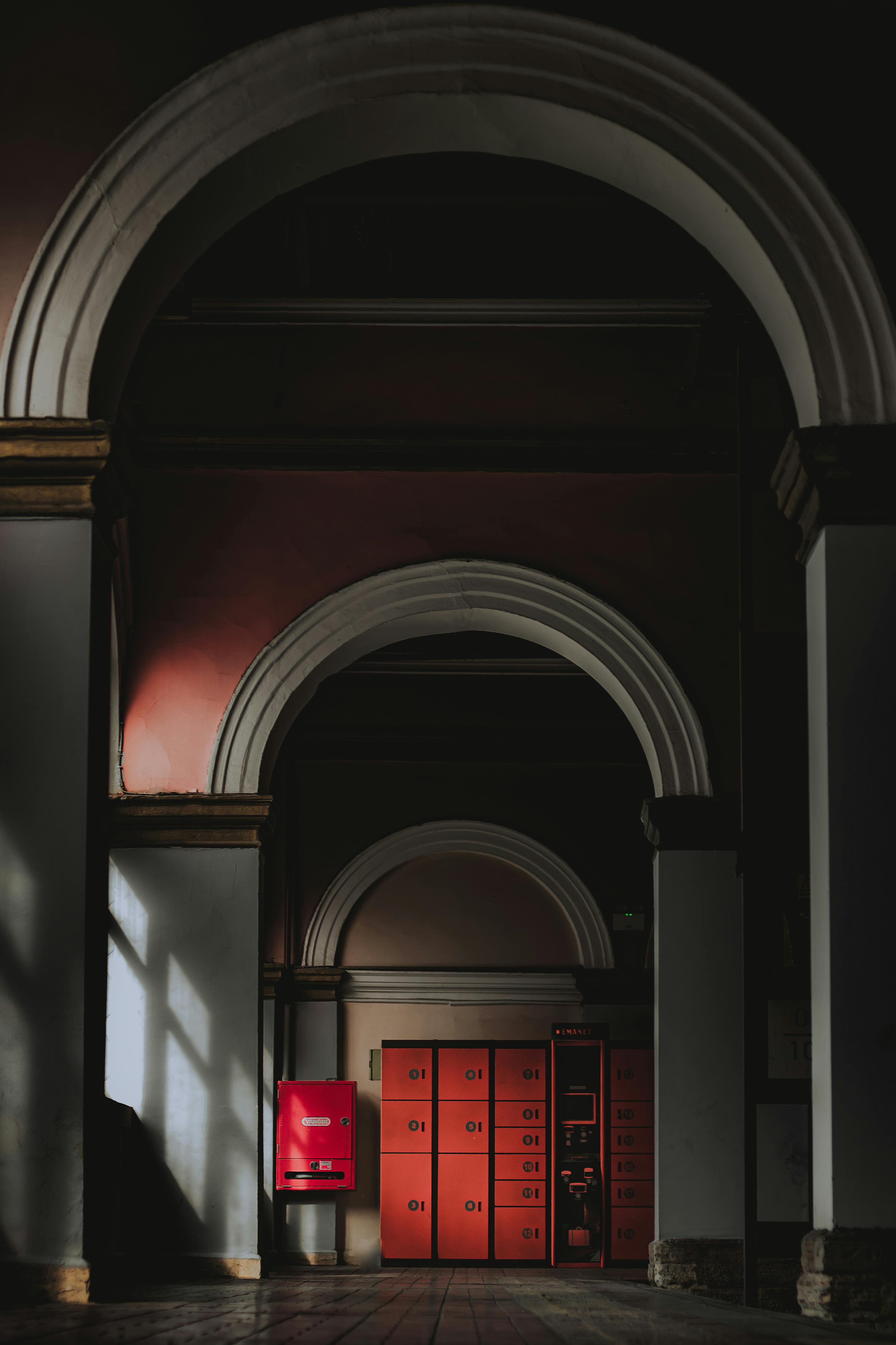 Dimly lit interior with white arches framing red lockers and a red fire alarm box on a tiled floor.