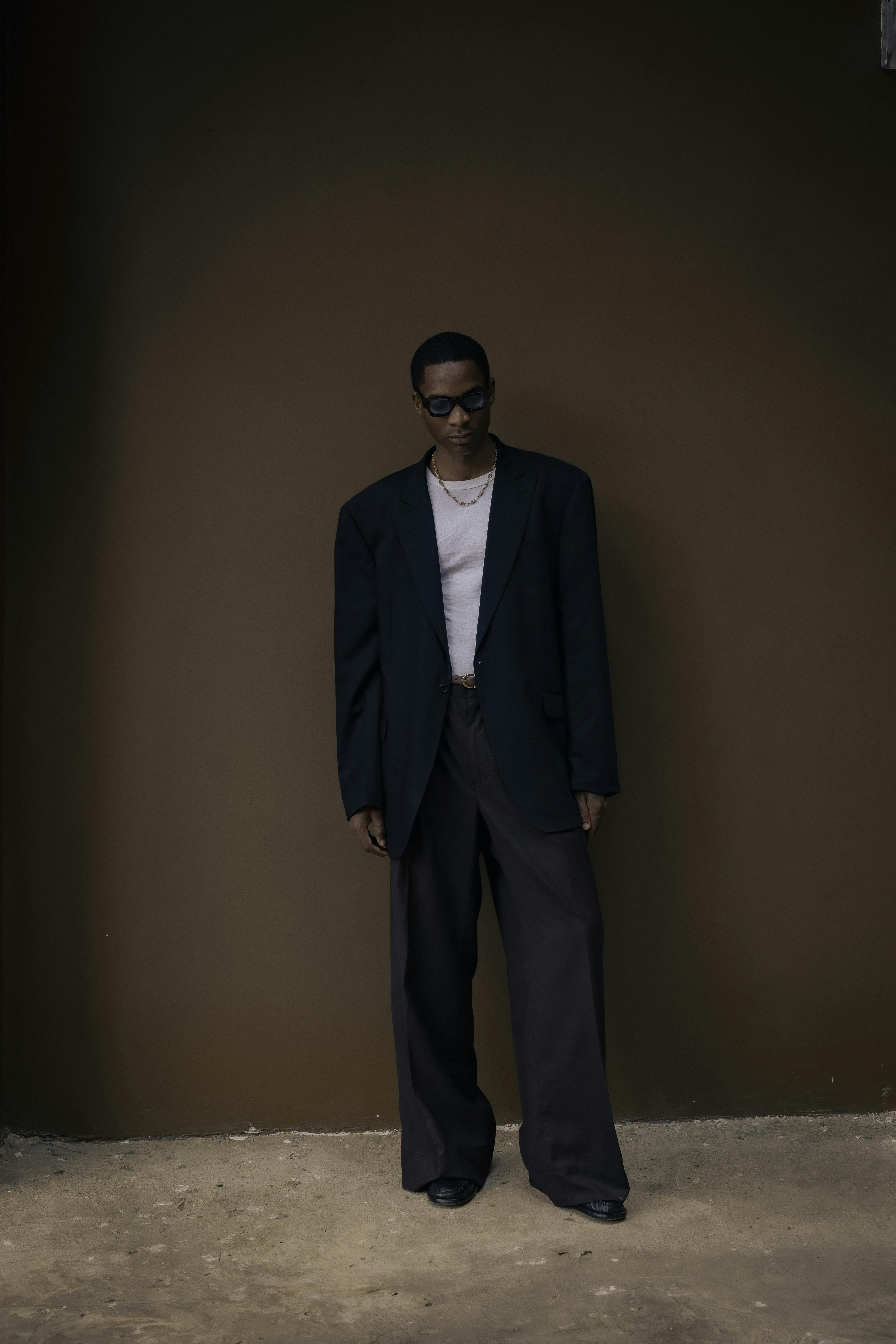 Man wearing oversized black blazer, wide-legged pants, sunglasses, and gold necklaces standing against a plain brown wall.