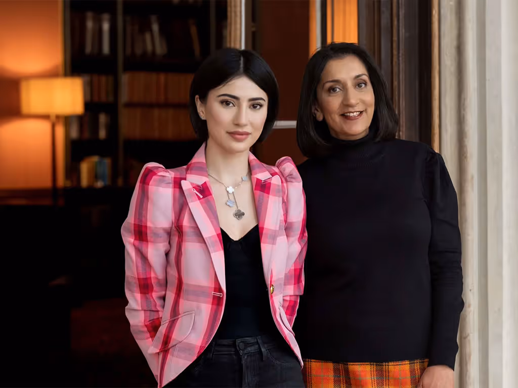 Two women standing indoors, one wearing a pink plaid blazer and black top, the other in a black top and orange plaid skirt.