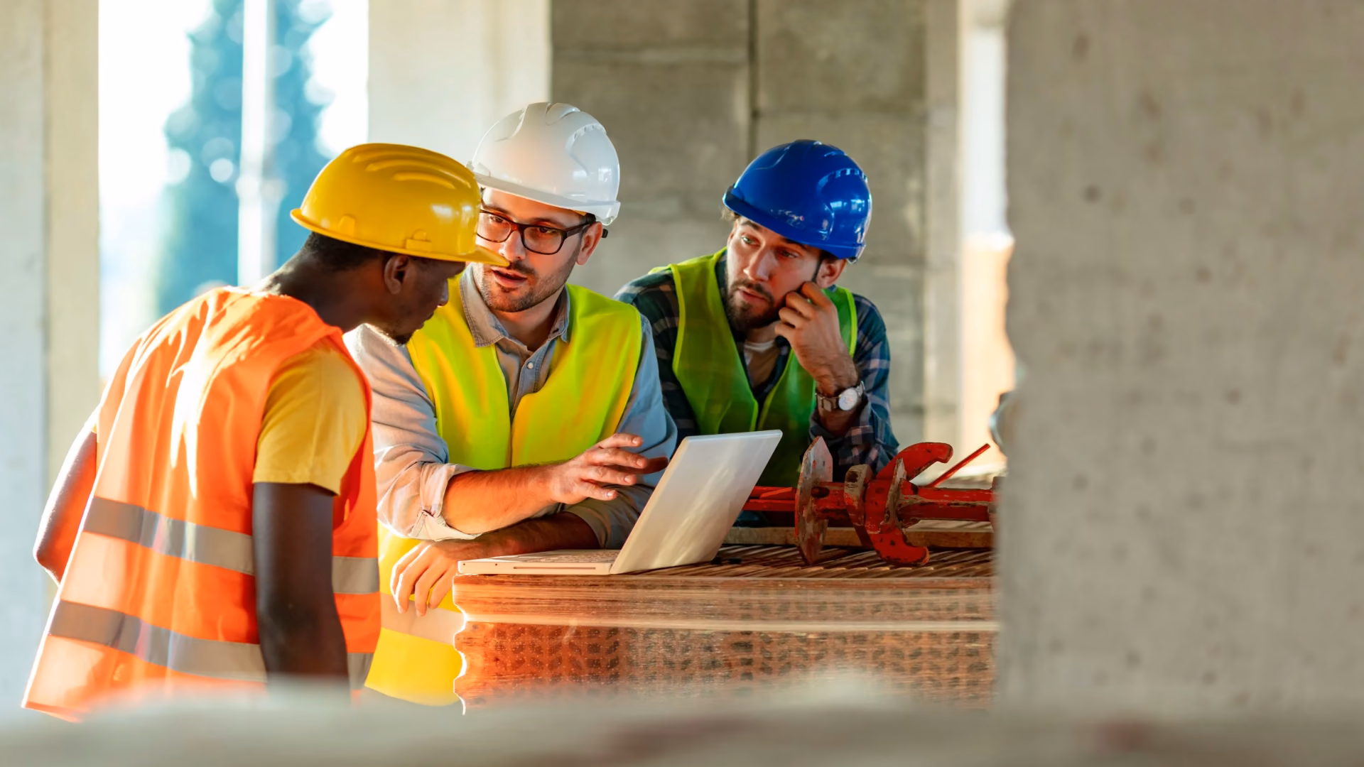 Three construction workers in hard hats and hi-vis vests reviewing a laptop on site