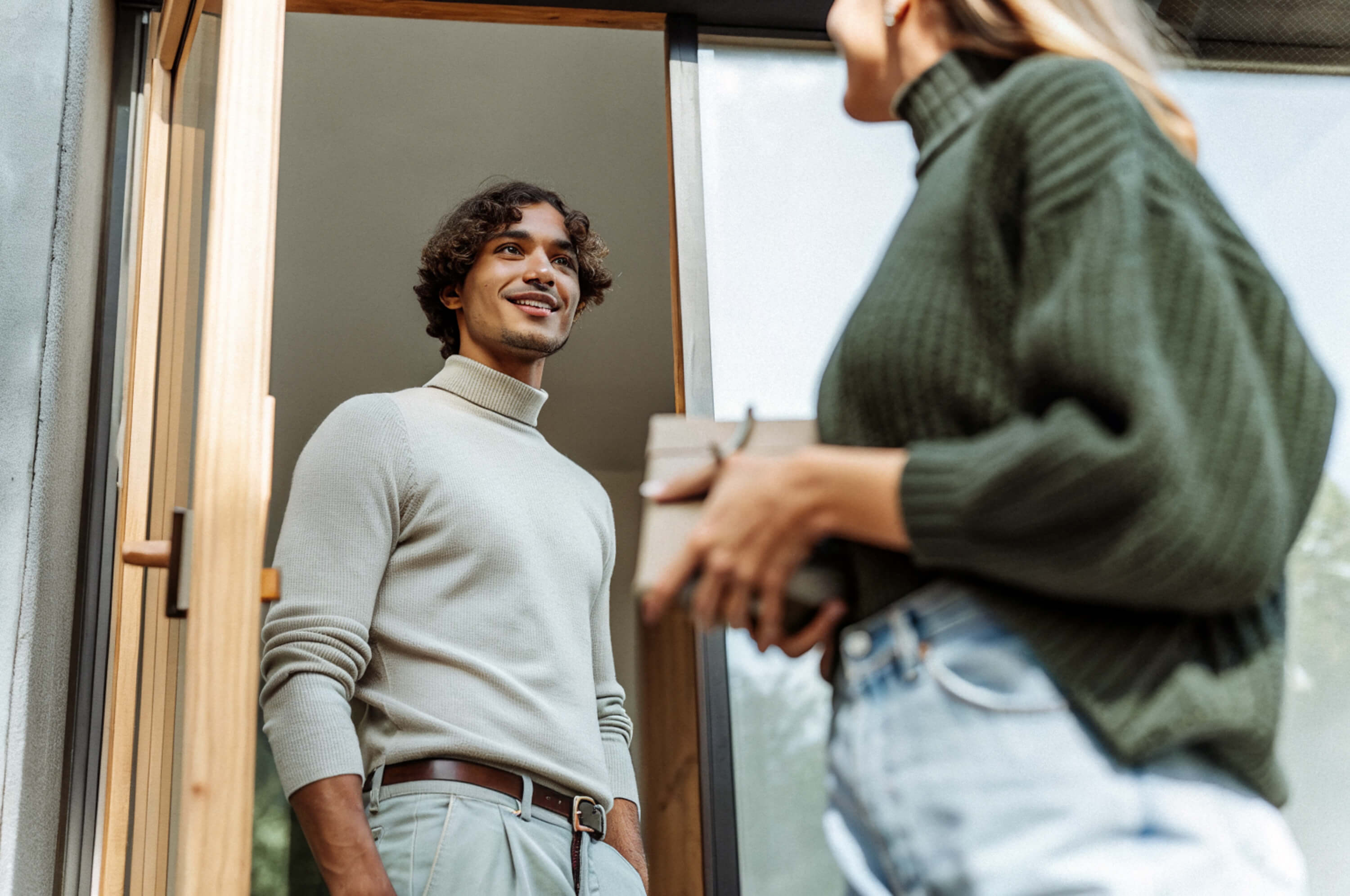 Man in a light sweater standing at a doorway smiling at a woman holding a small box.