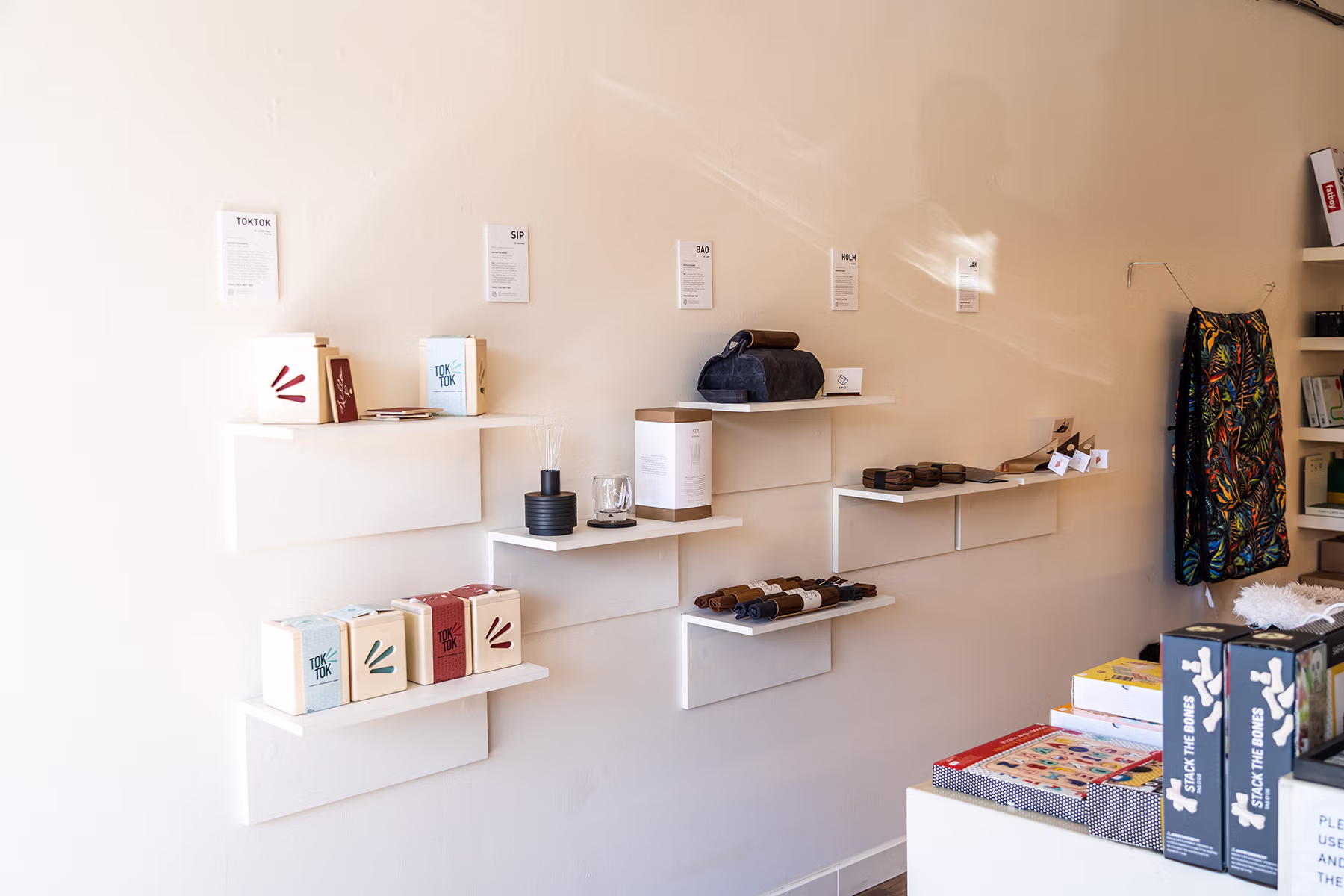 Interior retail display wall with minimalist white shelving holding designer lifestyle products. Items include wooden and fabric BAO rolls, boxed TOK TOK products, a SIP diffuser set, small wooden accessories, and printed product placards mounted above eac