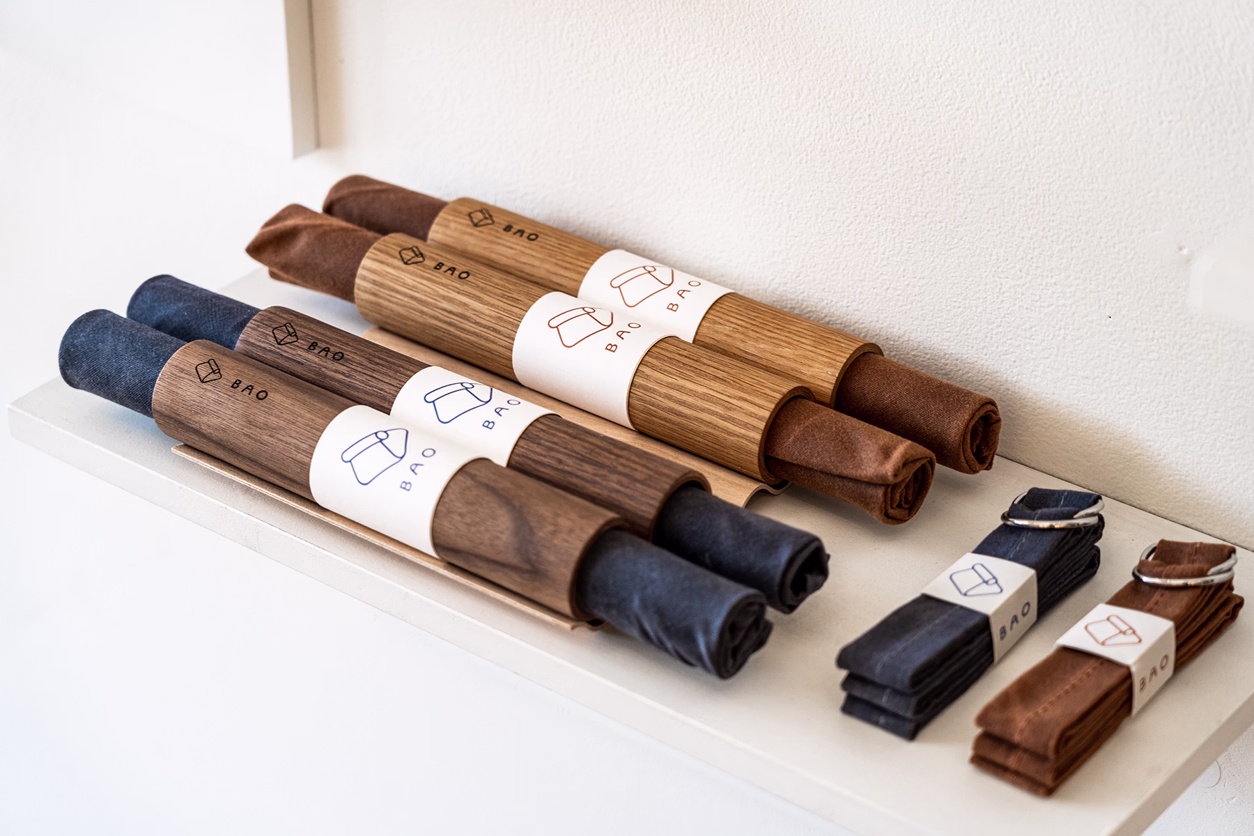 Close-up of multiple BAO bags made of wood and fabric arranged on a white display shelf. Each roll contains folded brown or dark charcoal fabric secured inside curved wooden handles with BAO-branded paper label wraps.