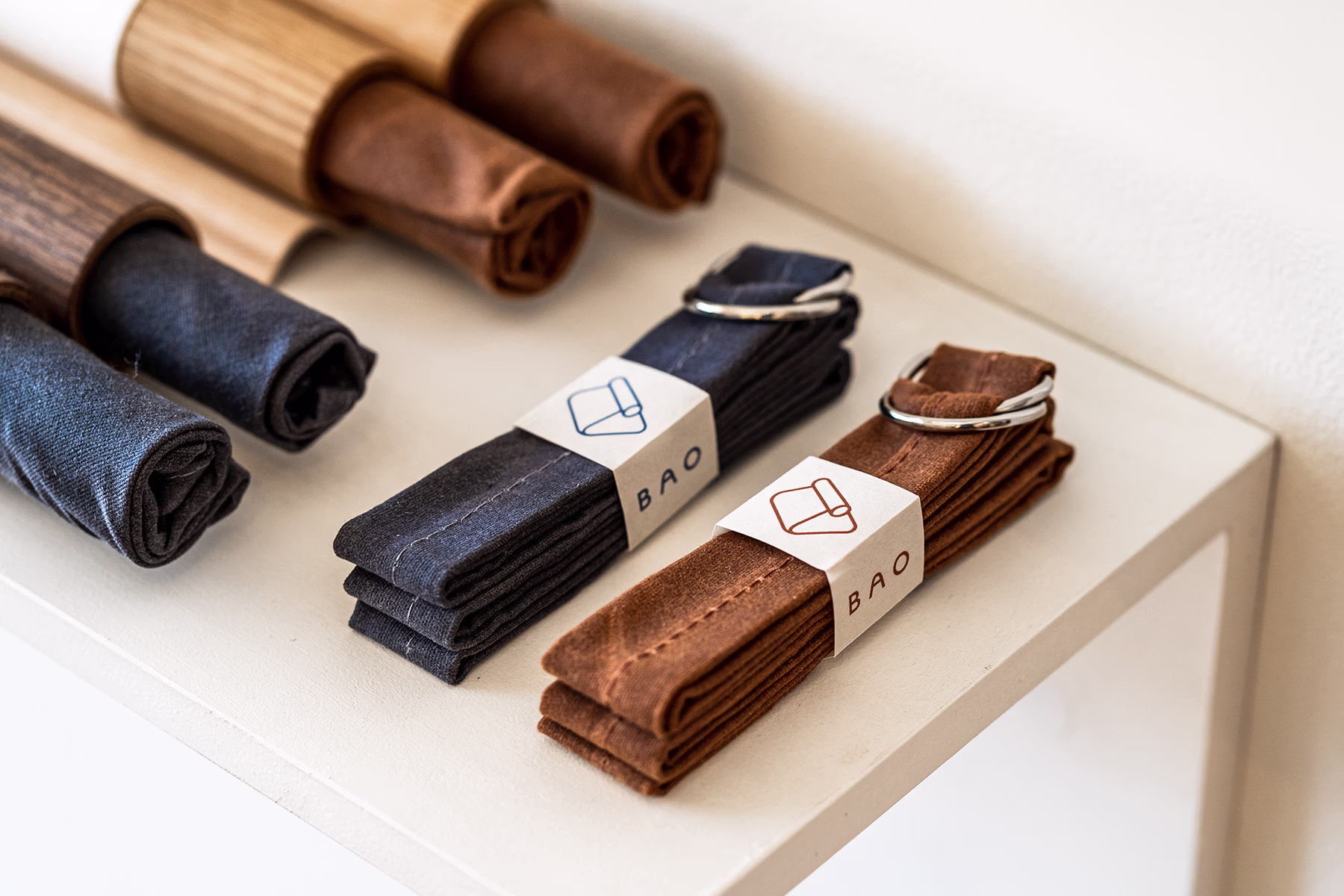 Close-up of folded BAO fabric straps in navy and brown, each secured with metal D-rings and wrapped with branded paper labels, arranged neatly on a white display shelf.