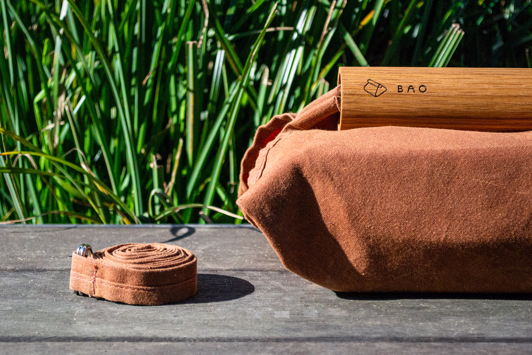 A rolled brown strap and a brown fabric bundle with a light wooden BAO handle sit on a wooden bench outdoors, with tall green grass blurred in the background.