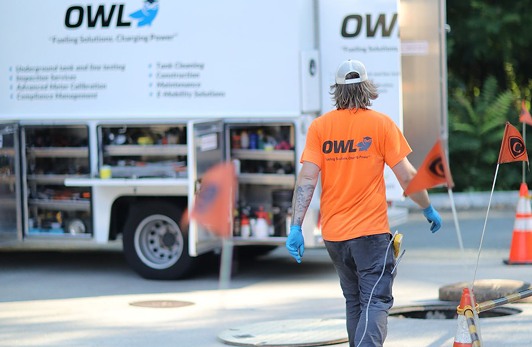 Worker wearing an orange OWL t-shirt and blue gloves walking near utility truck and safety cones on the street.