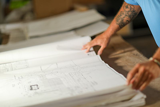 Person with tattoos reviewing architectural blueprints on a wooden table.