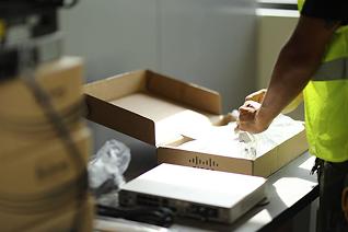 Worker in a yellow safety vest packing an item in a cardboard box on a table with a scale nearby.