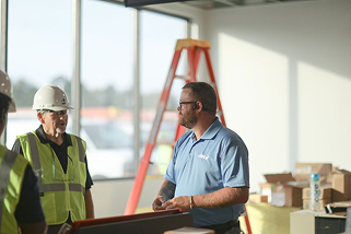 Two construction professionals talking indoors, one wearing a safety vest and helmet, with orange ladder and boxes in the background.