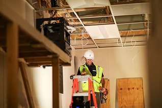 Worker in orange shirt and cap inspecting a manhole on a street with traffic cones in the background.