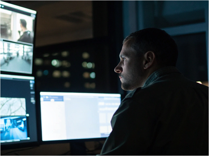 Man in a dark room monitoring multiple screens showing surveillance footage.
