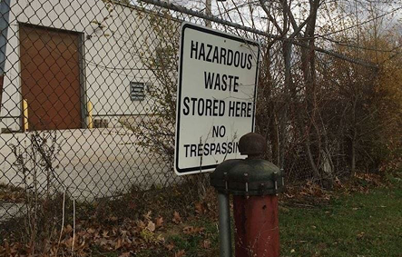 Chain-link fence with a sign reading 'Hazardous Waste Stored Here No Trespassing' near a red fire hydrant.