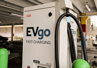 Person in orange shirt and blue gloves working on pipes under a fuel dispenser with pipes and tools nearby.
