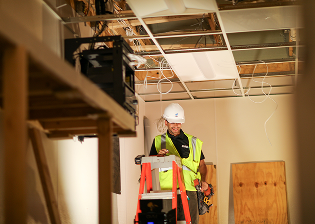 Person wearing a white helmet and neon green safety vest installing a security camera onto a ceiling.