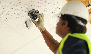 Maintenance worker in safety gear installing or inspecting a security camera on a ceiling.