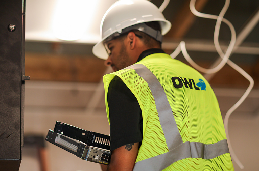 Technician wearing a white hard hat and gloves installing a security camera on a ceiling.