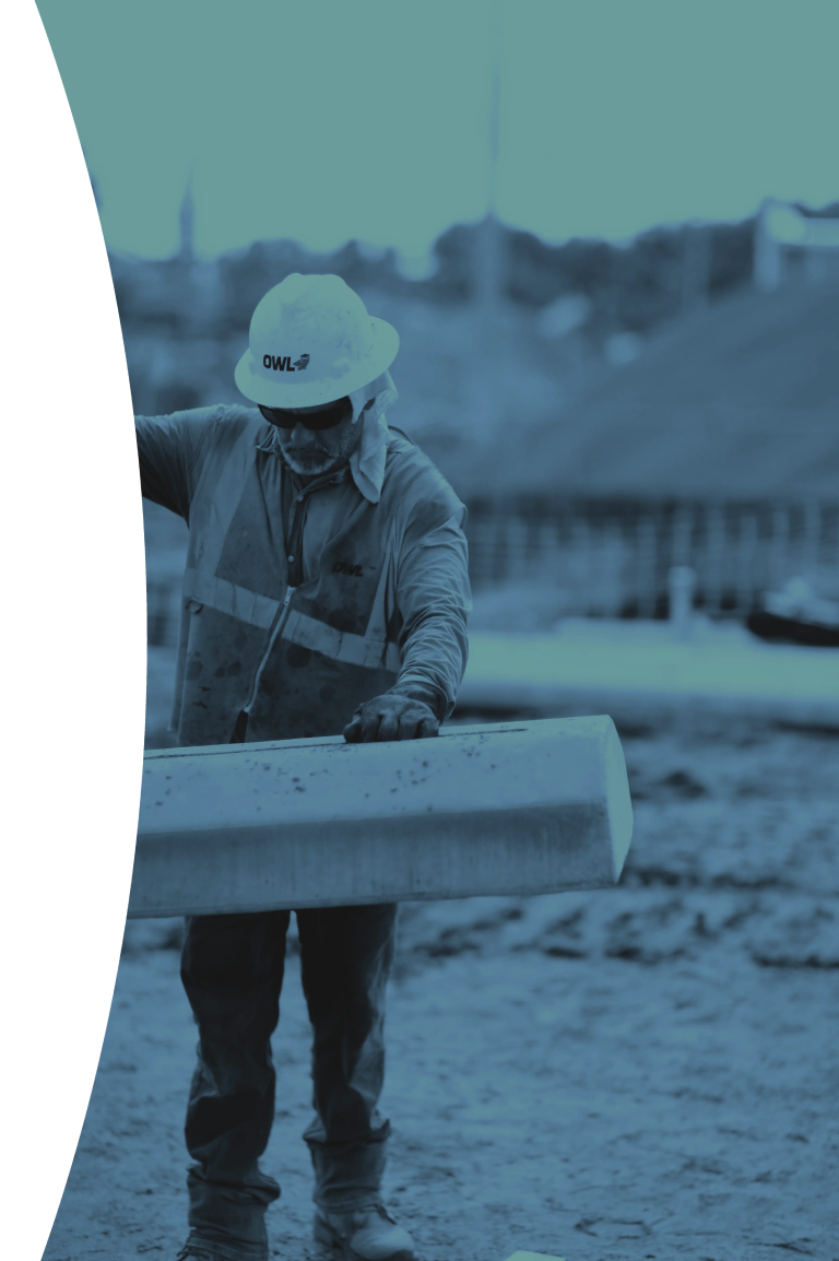 Construction worker wearing a hard hat and safety vest examining a concrete "deadman" beam on a job site.