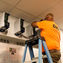 Technician in orange shirt using a screwdriver to work on an electrical panel while standing on a blue ladder.