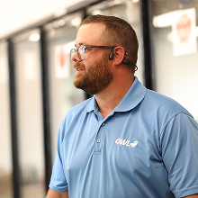 Man with glasses and beard wearing a light blue polo shirt and Bluetooth earpiece looking to the left in a bright indoor setting.