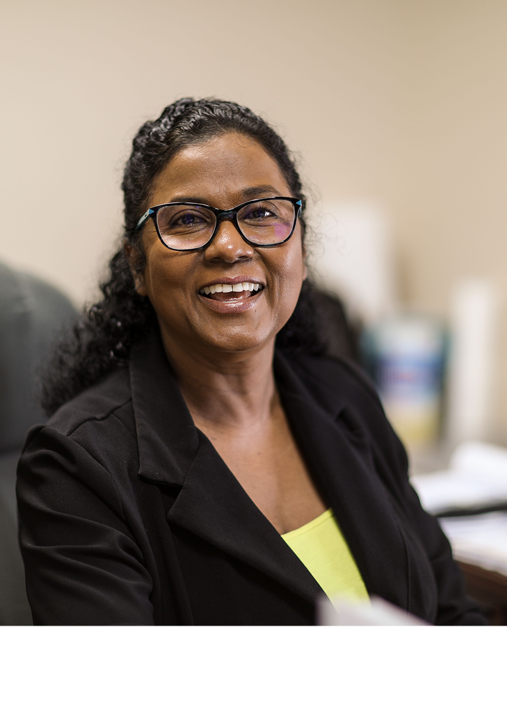 Smiling woman with curly hair and glasses wearing a black blazer and yellow top in an office setting.